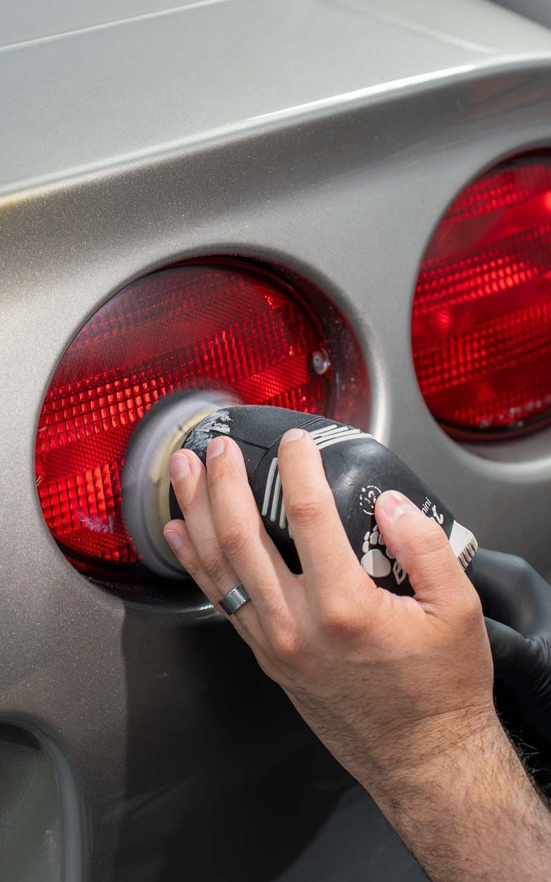 A hand polishes a car tail light with a rotating buffer, likely to restore clarity.