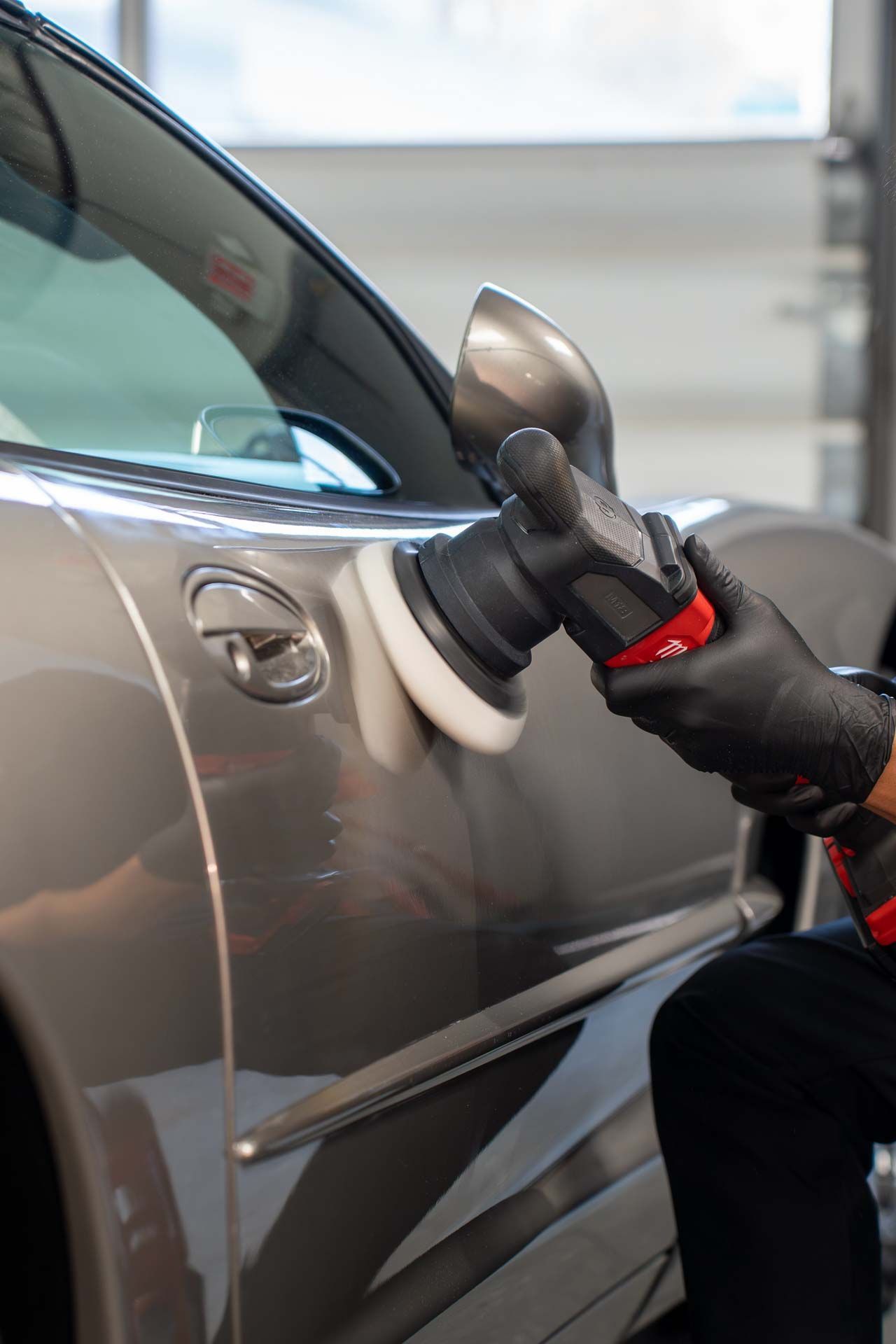 Person polishes a gray car door with a buffing machine, wearing black gloves.