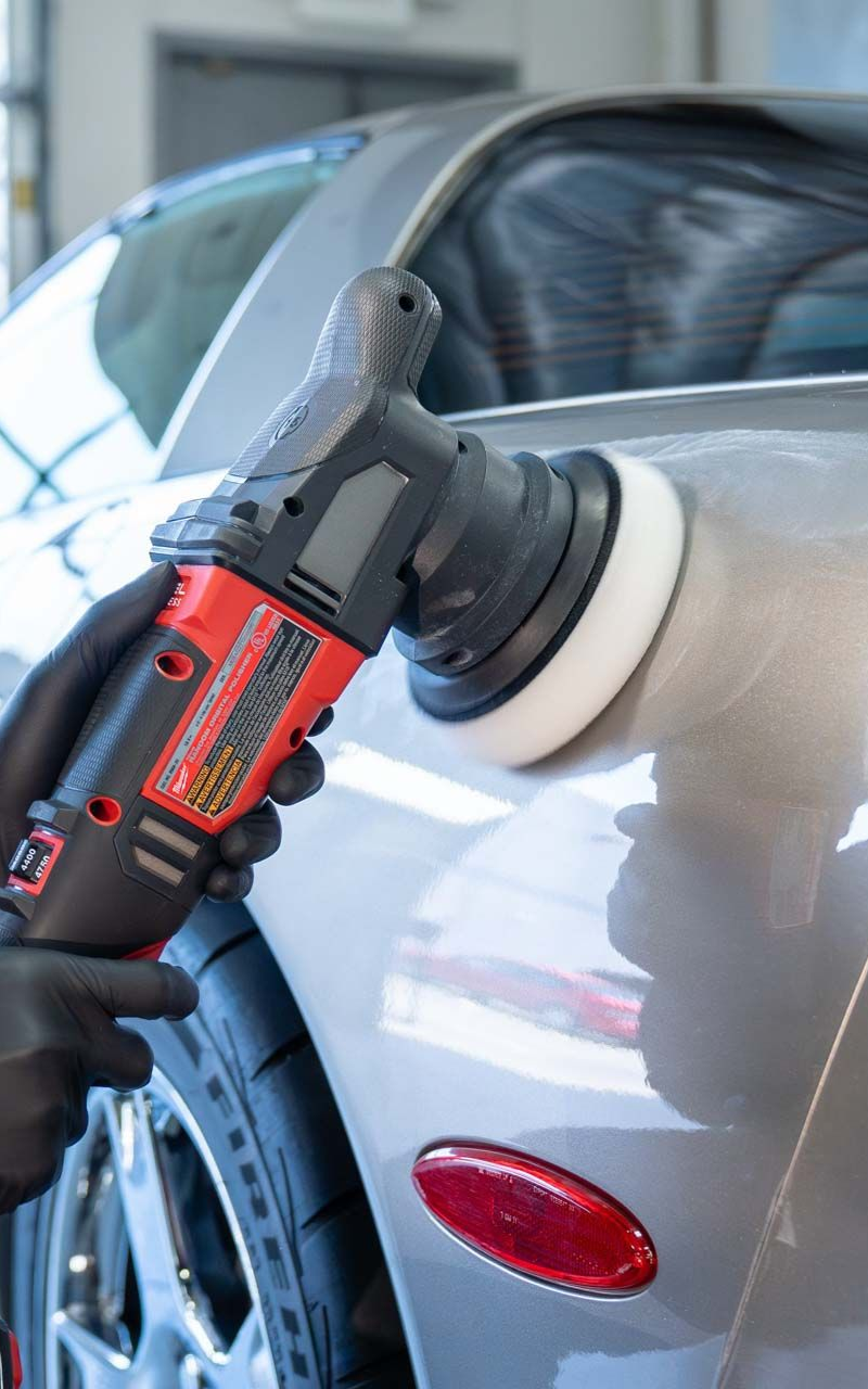 Person in black gloves polishes a silver car with a red and black power buffer in a garage.