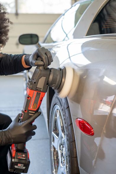 Person in gloves applying a detailing product to a car's gray hood with a spray bottle and applicator.