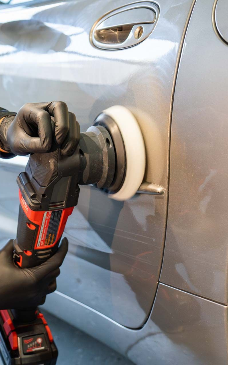 Person using a power polisher on a silver car door. Black gloves, red and black tool, white buffing pad.