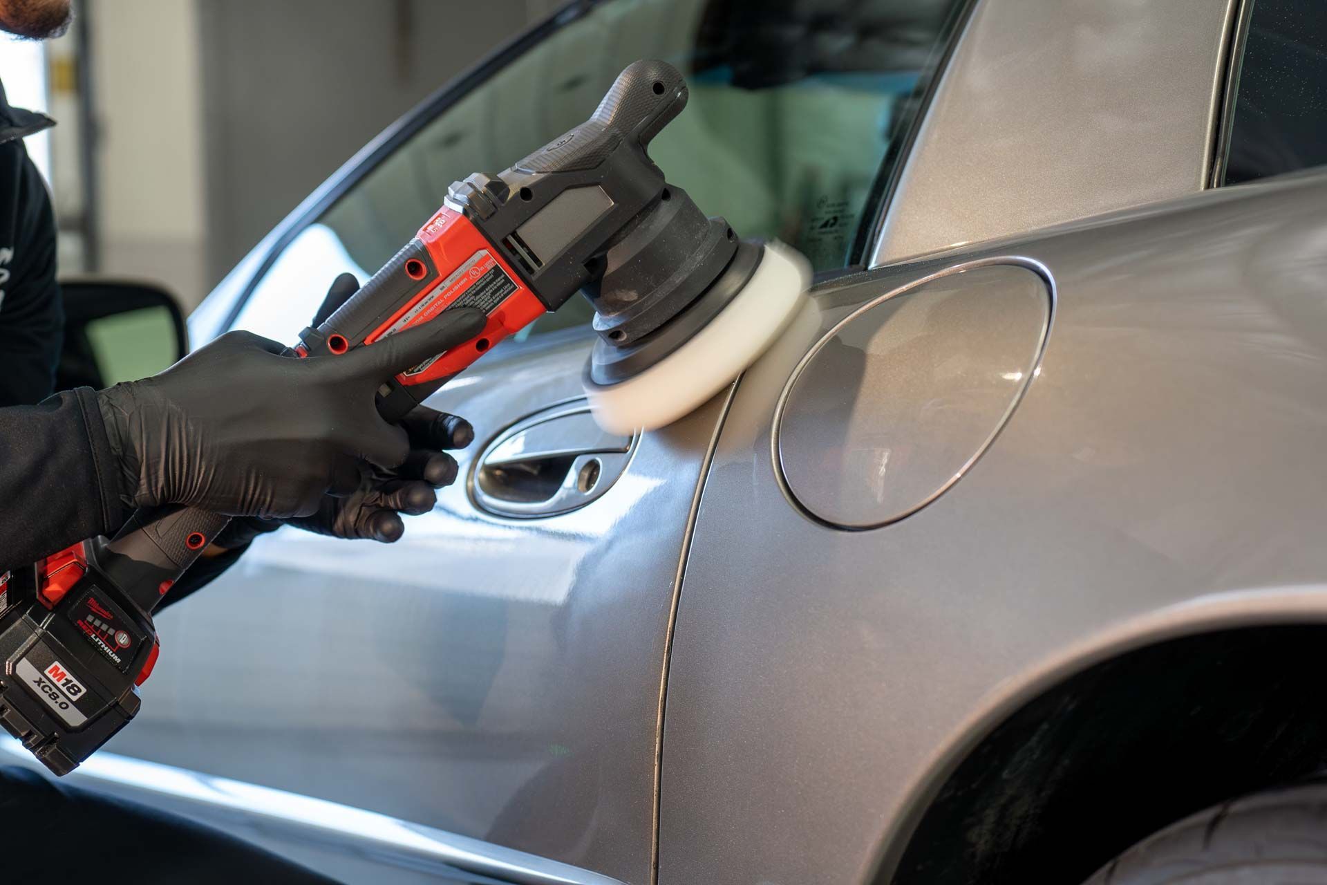 Person using an electric polisher on a silver car's gas tank door, wearing black gloves.