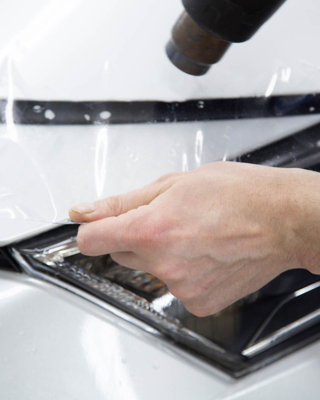 A hand holds clear film, applying it to a car part; heat gun is directed towards it.