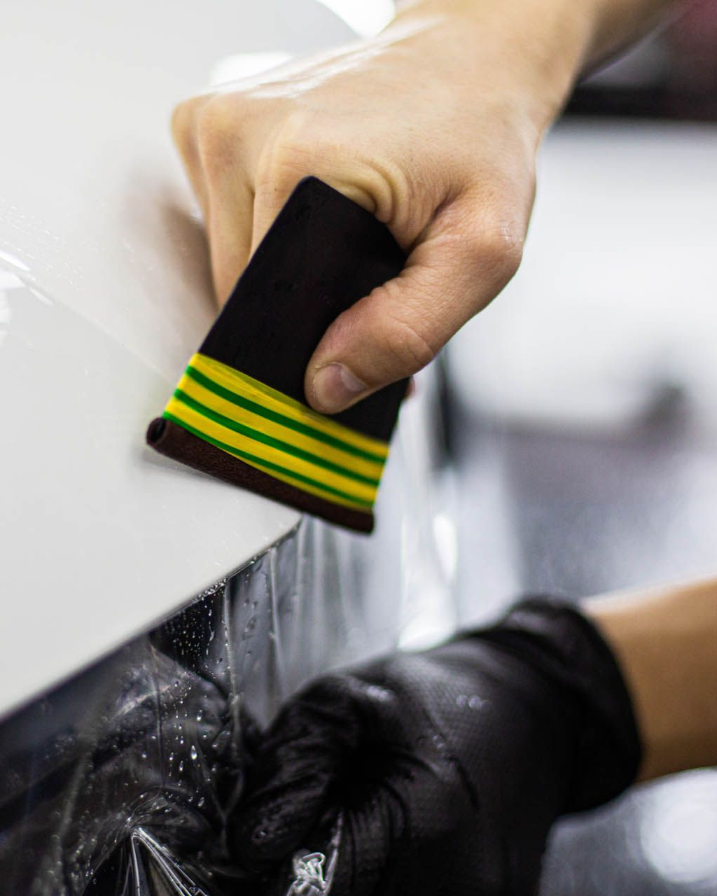 Hand applying film to a white surface using a smoothing tool, other hand wearing a black glove.