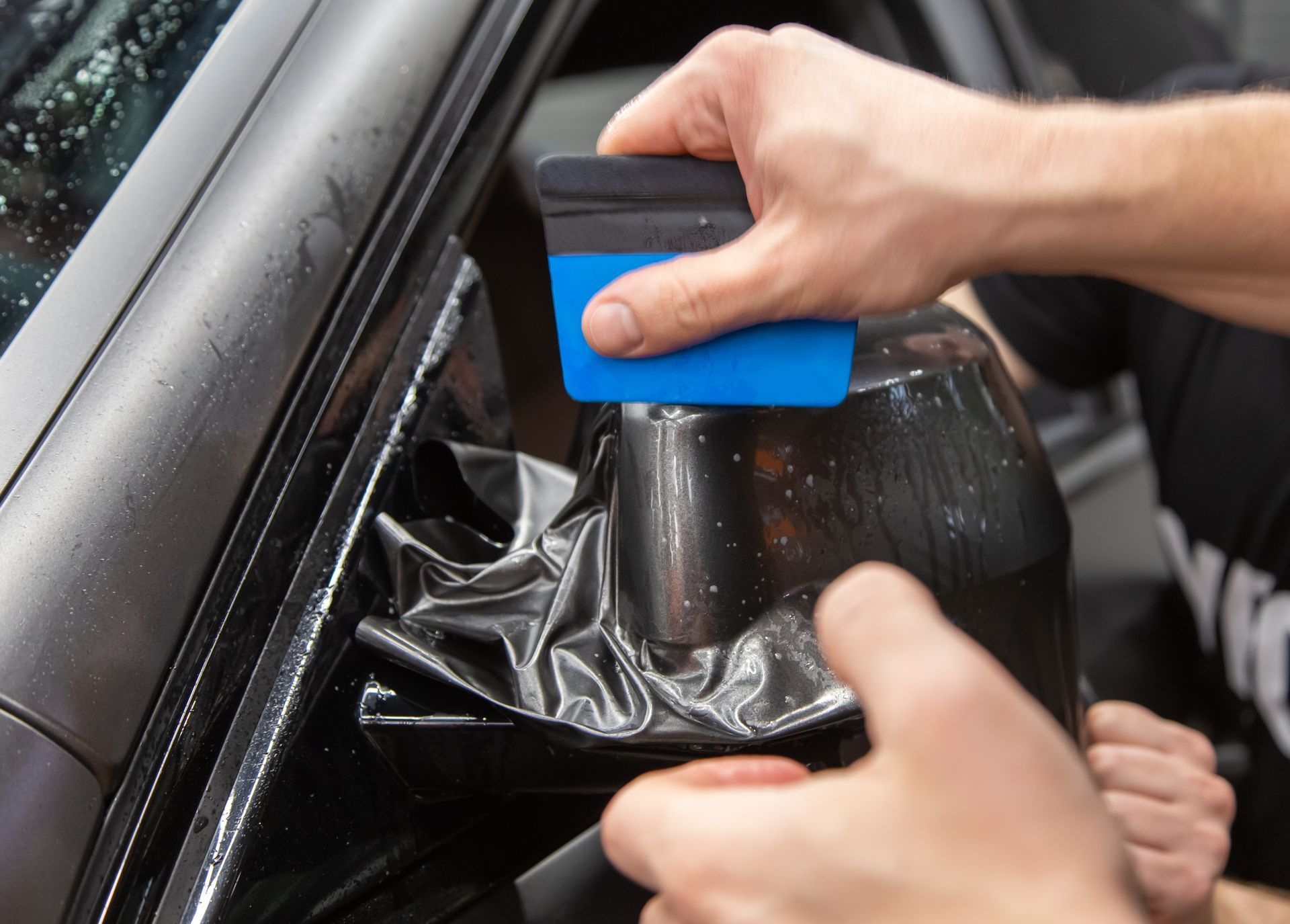 Person applying black vinyl wrap to a car's side mirror with a blue squeegee.