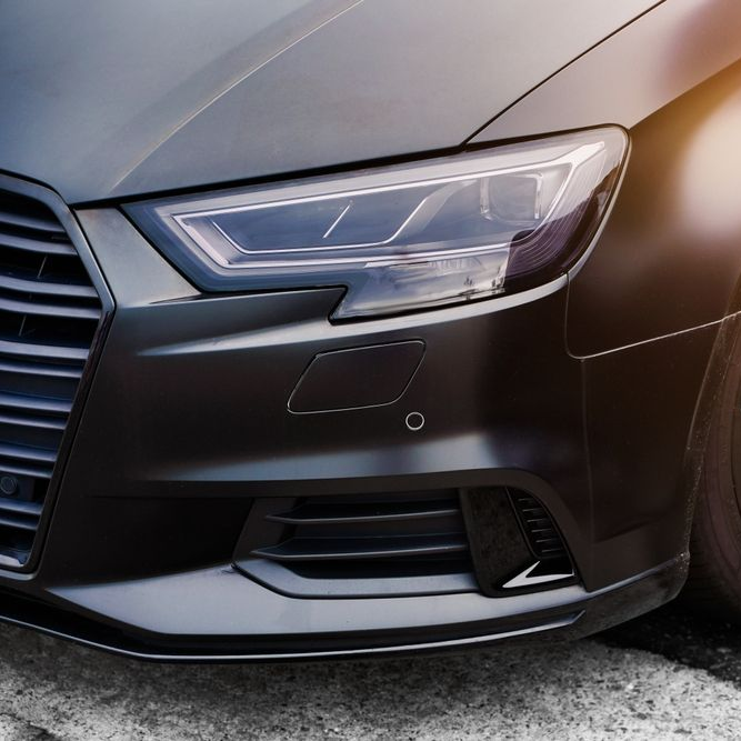 Black car front, focus on headlight and bumper, parked on concrete.