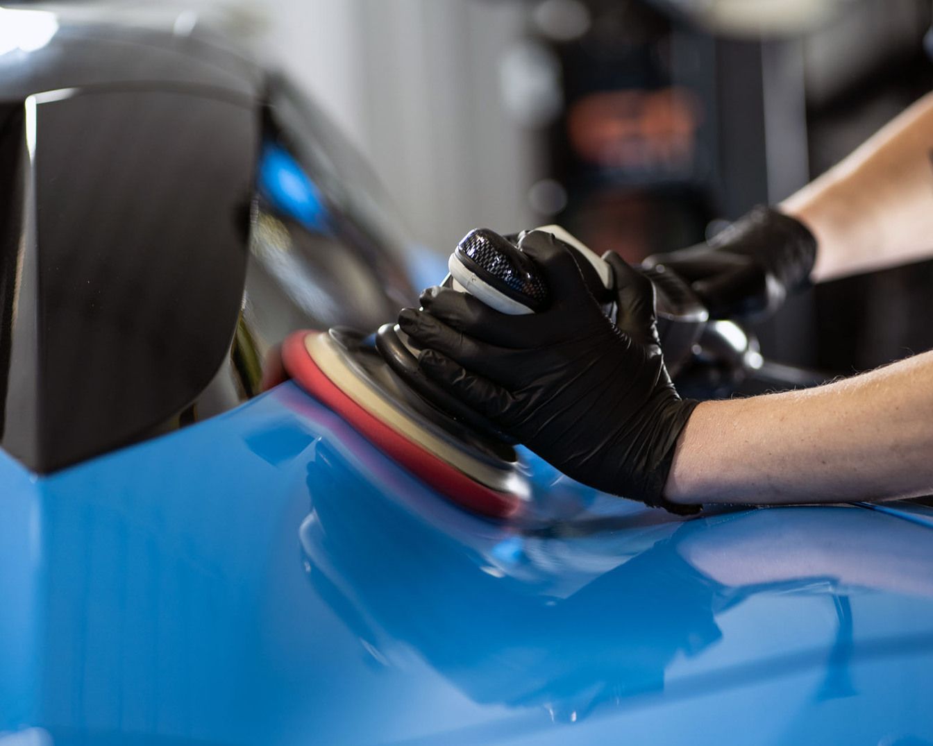 Person polishes a white car bumper with a buffing machine, indoors.