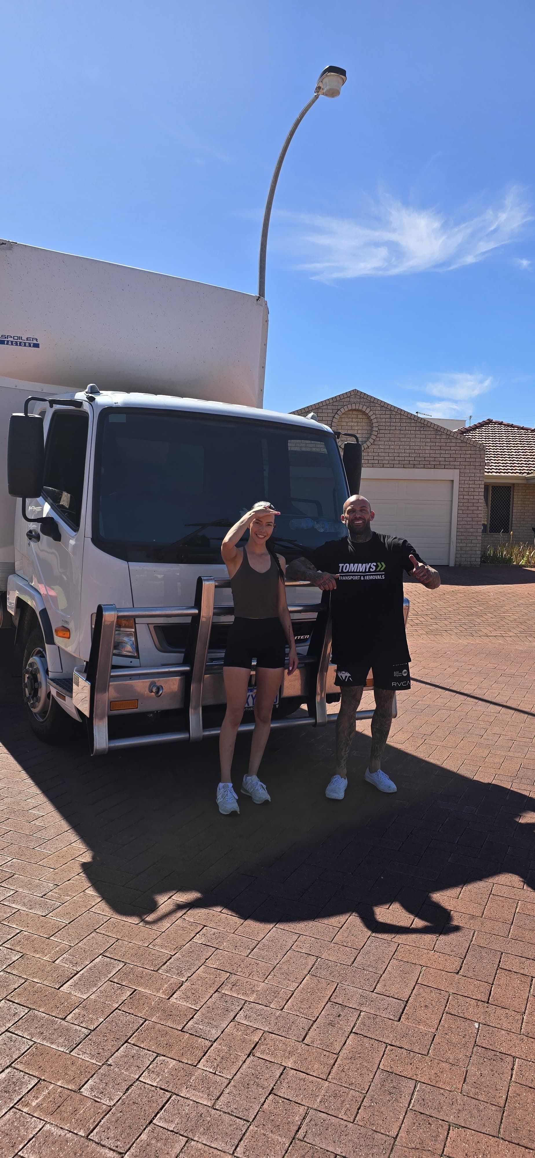 Two people stand near a white truck on a sunny day. A curved street light is overhead.