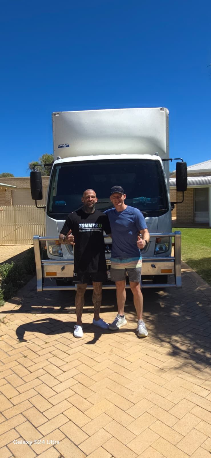Two men stand in front of a box truck on a brick driveway, posing with thumbs up.
