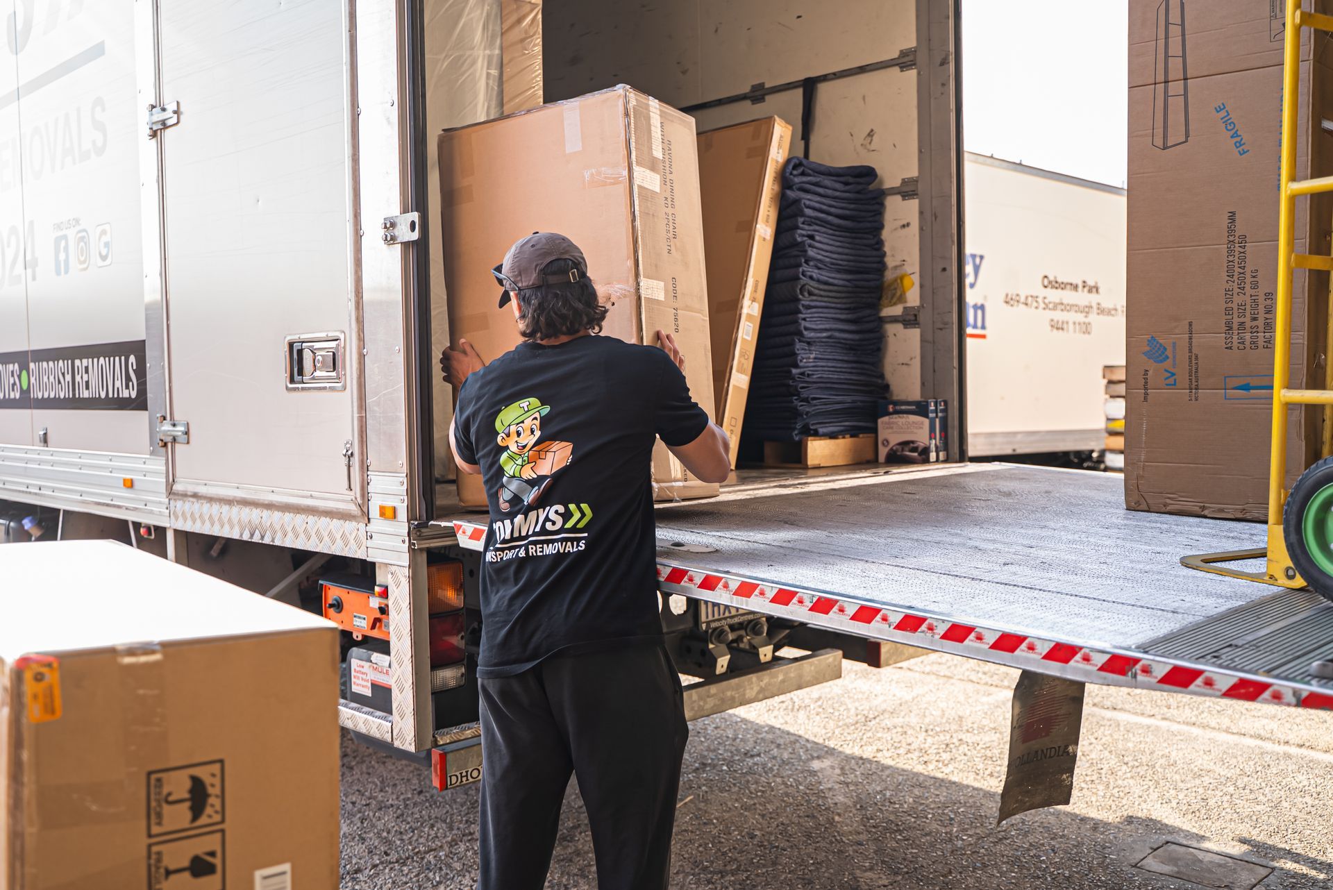 A removalist from Tommy's Transport and Removals unloads a large cardboard box from back of a parked moving truck.