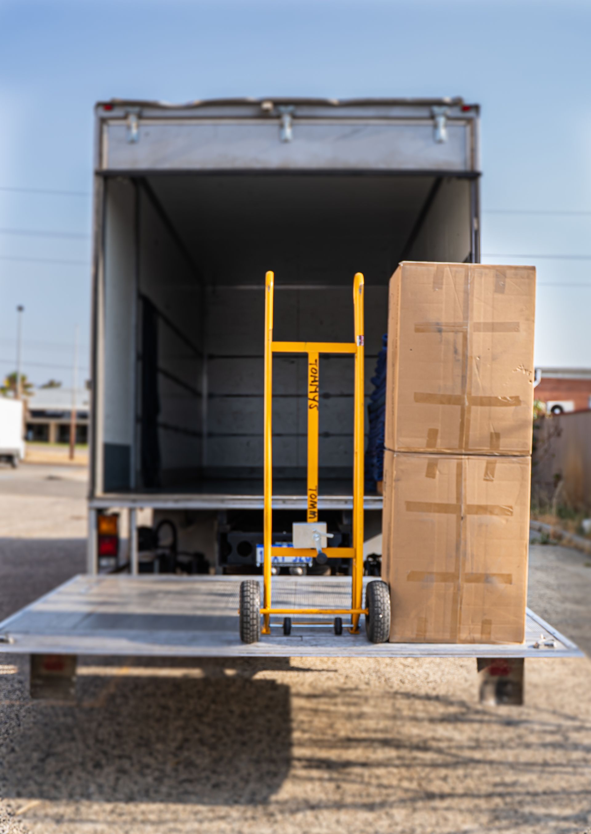 A yellow hand truck stands on a truck lift gate, next to a large cardboard box, in front of an open moving truck.