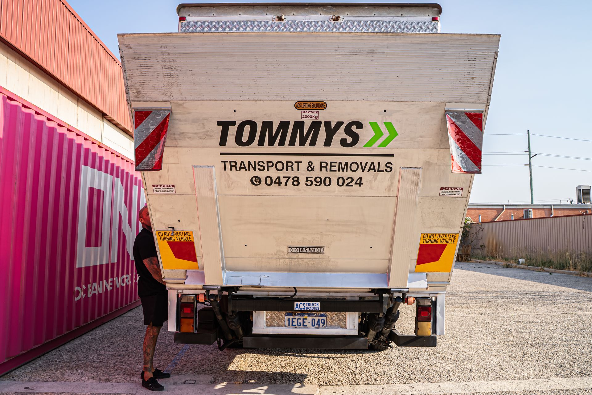 A person stands beside a Tommy’s Transport & Removals truck parked outdoors near a large pink storage container.