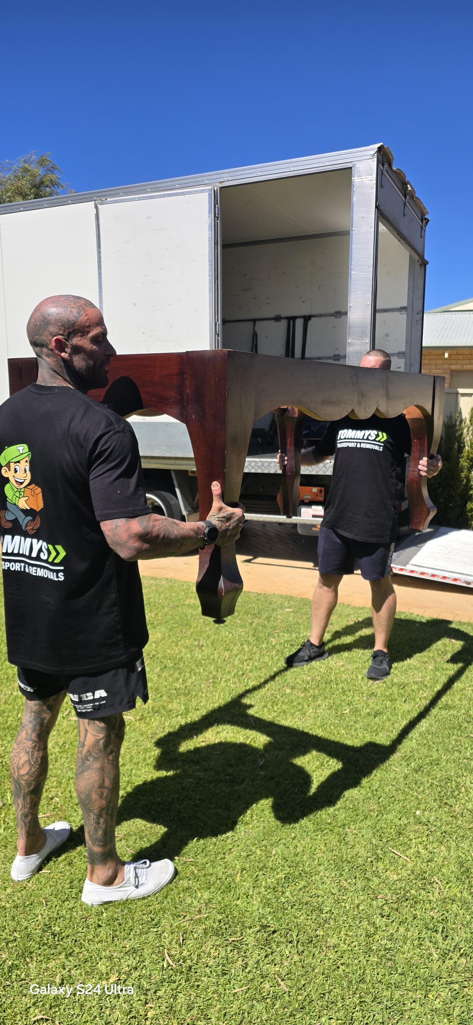 Two men carrying a large wooden object outdoors on a sunny day.