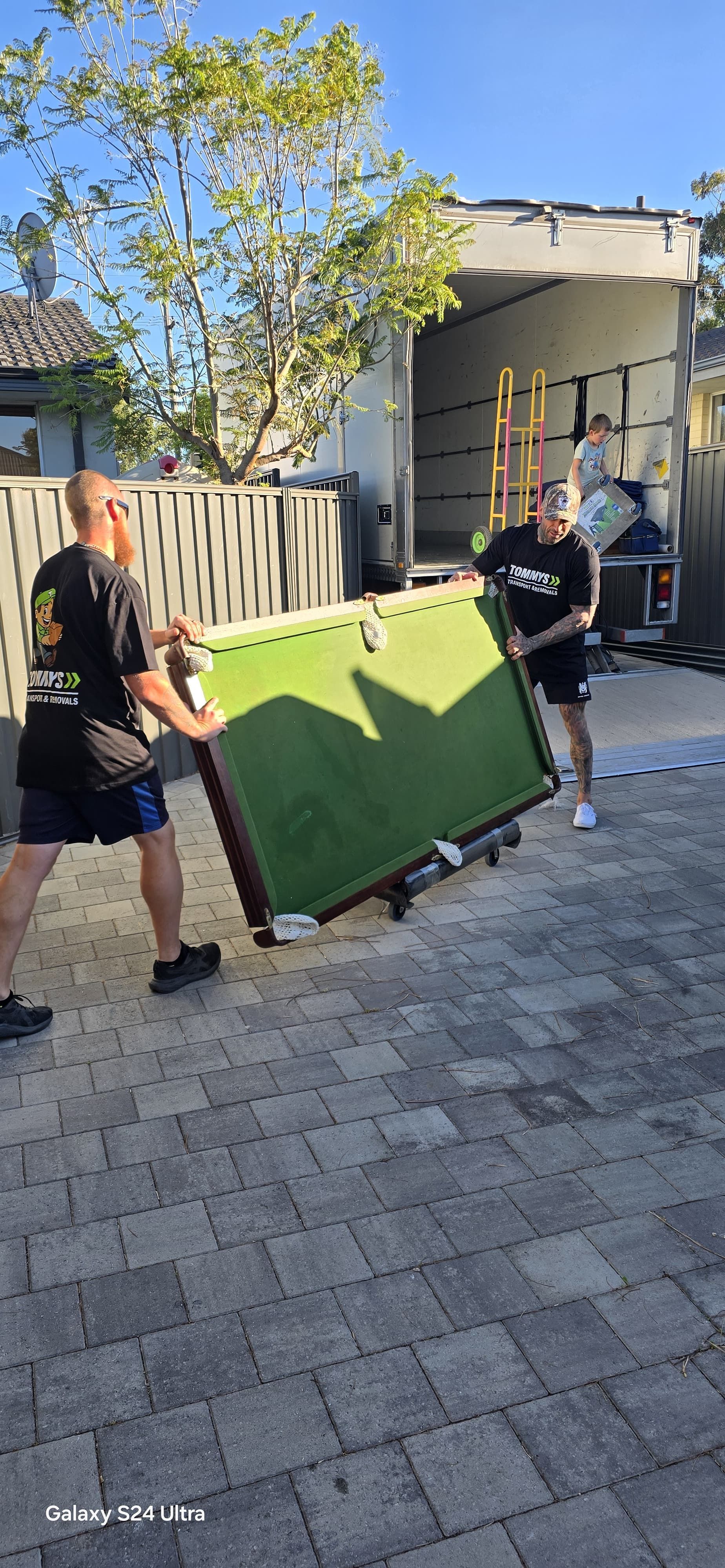Two people carry a green pool table toward the back of a moving truck on a sunny day.
