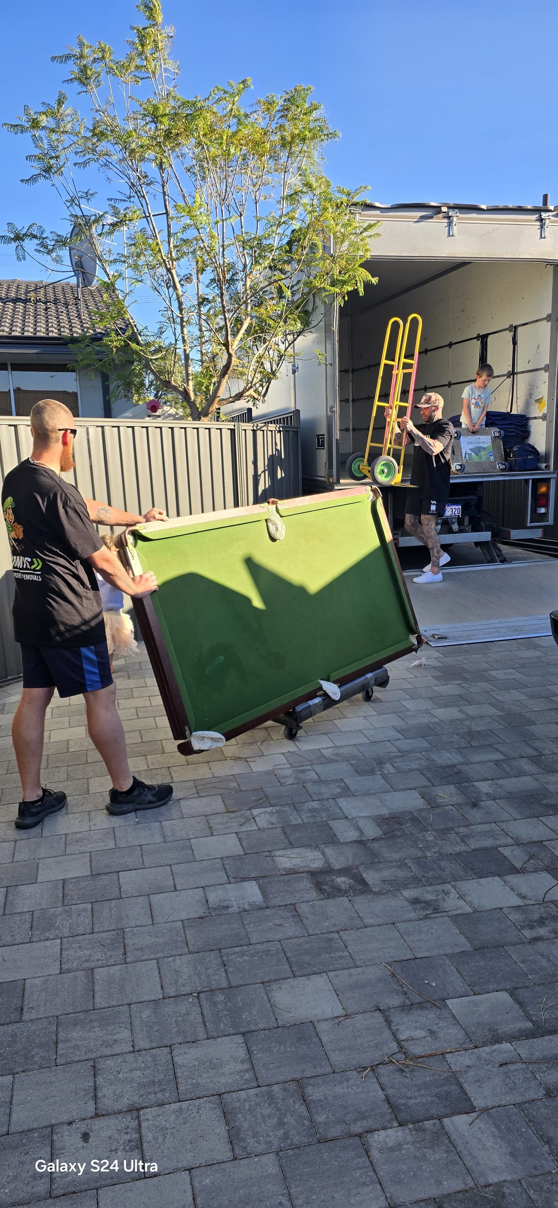 Person moving a green bench in a sunny backyard patio with a tree and open garage area