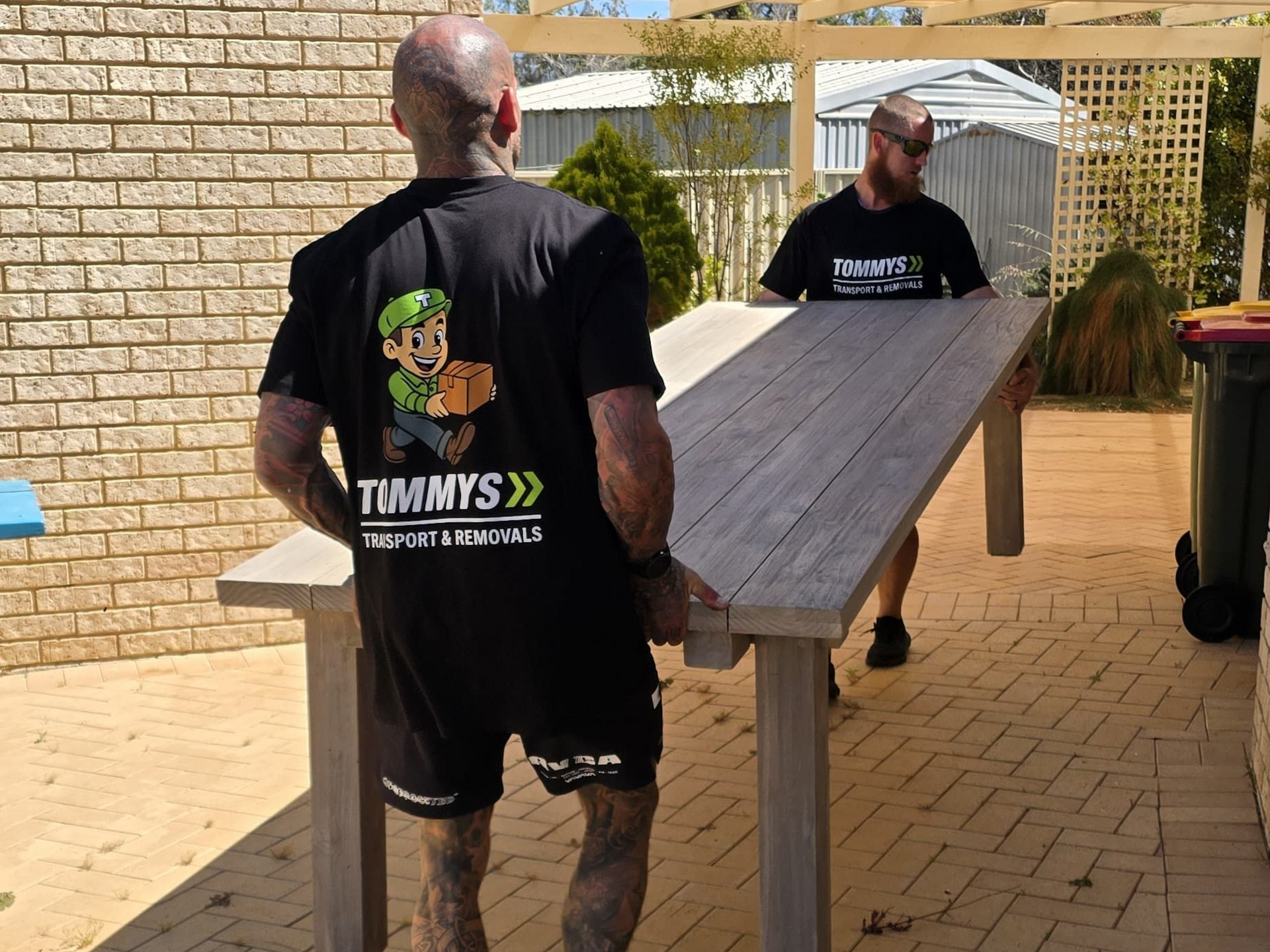 Two men in black shirts lift a wooden table outside a brick building.