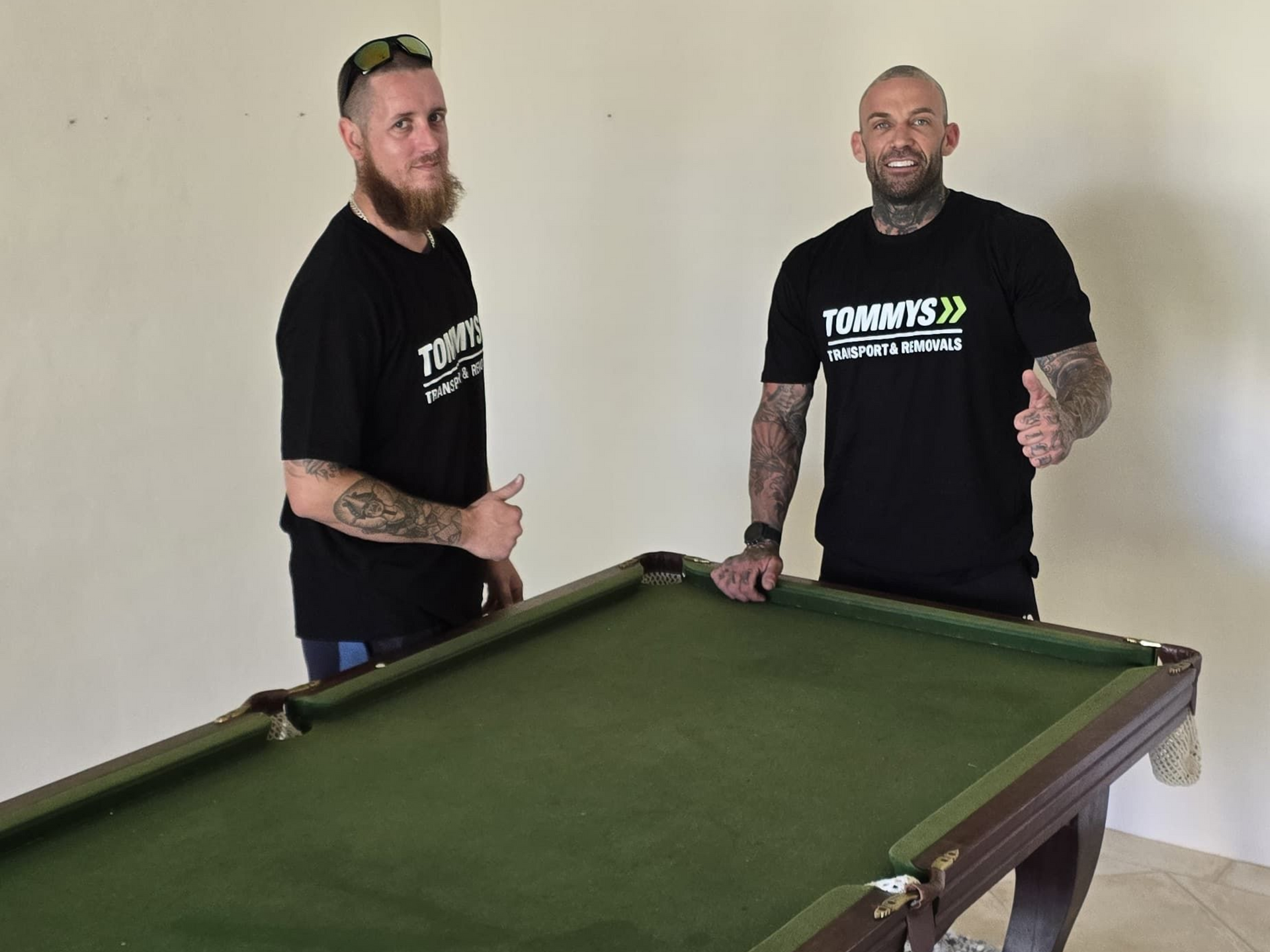 Two men standing behind a green pool table, both wearing shirts with Tommy's Transport and Removals logo, giving thumbs up.