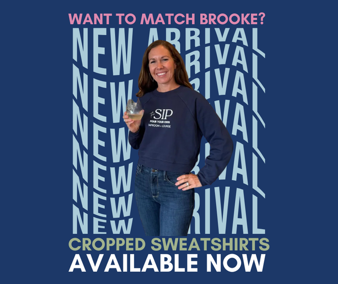 Woman in blue sweatshirt and jeans at wine tasting station, smiling, holding a glass.