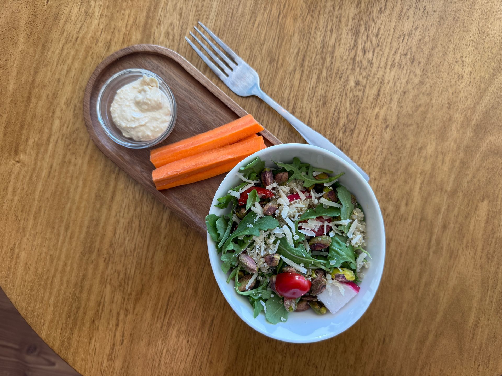 Salad with hummus, carrot sticks, and a fork on a wooden tray.