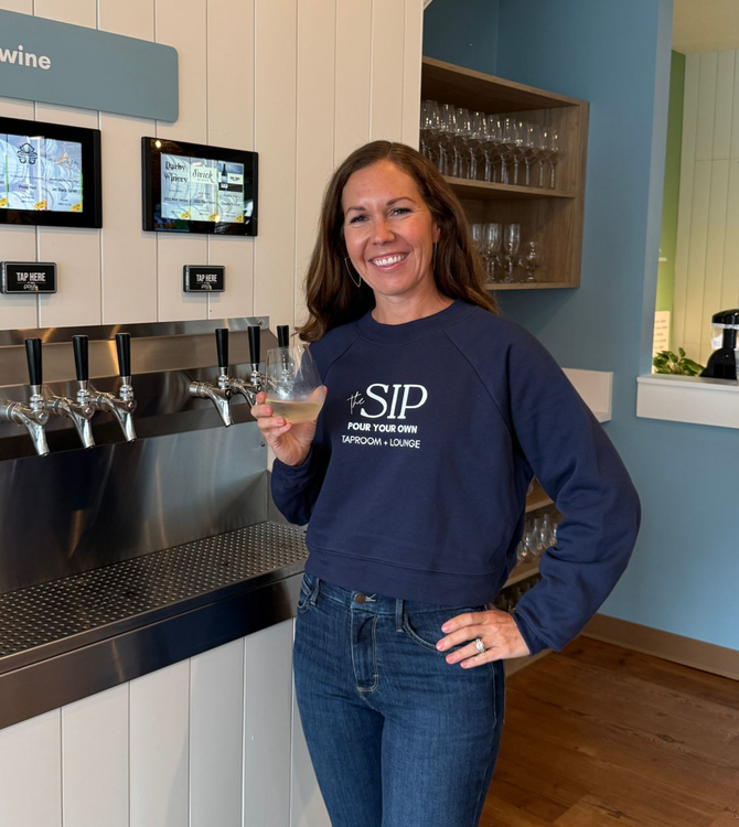 Woman in blue sweatshirt and jeans at wine tasting station, smiling, holding a glass.