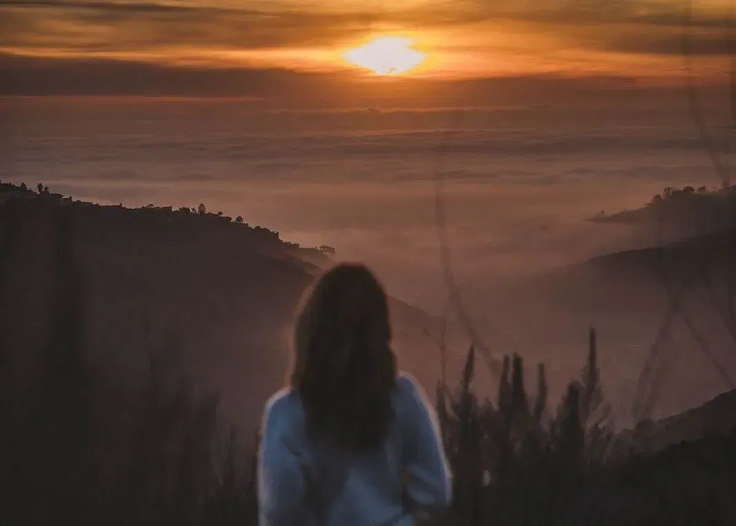 Woman overlooking mountain landscape at sunrise. Orange sky, fog, and silhouettes of hills.