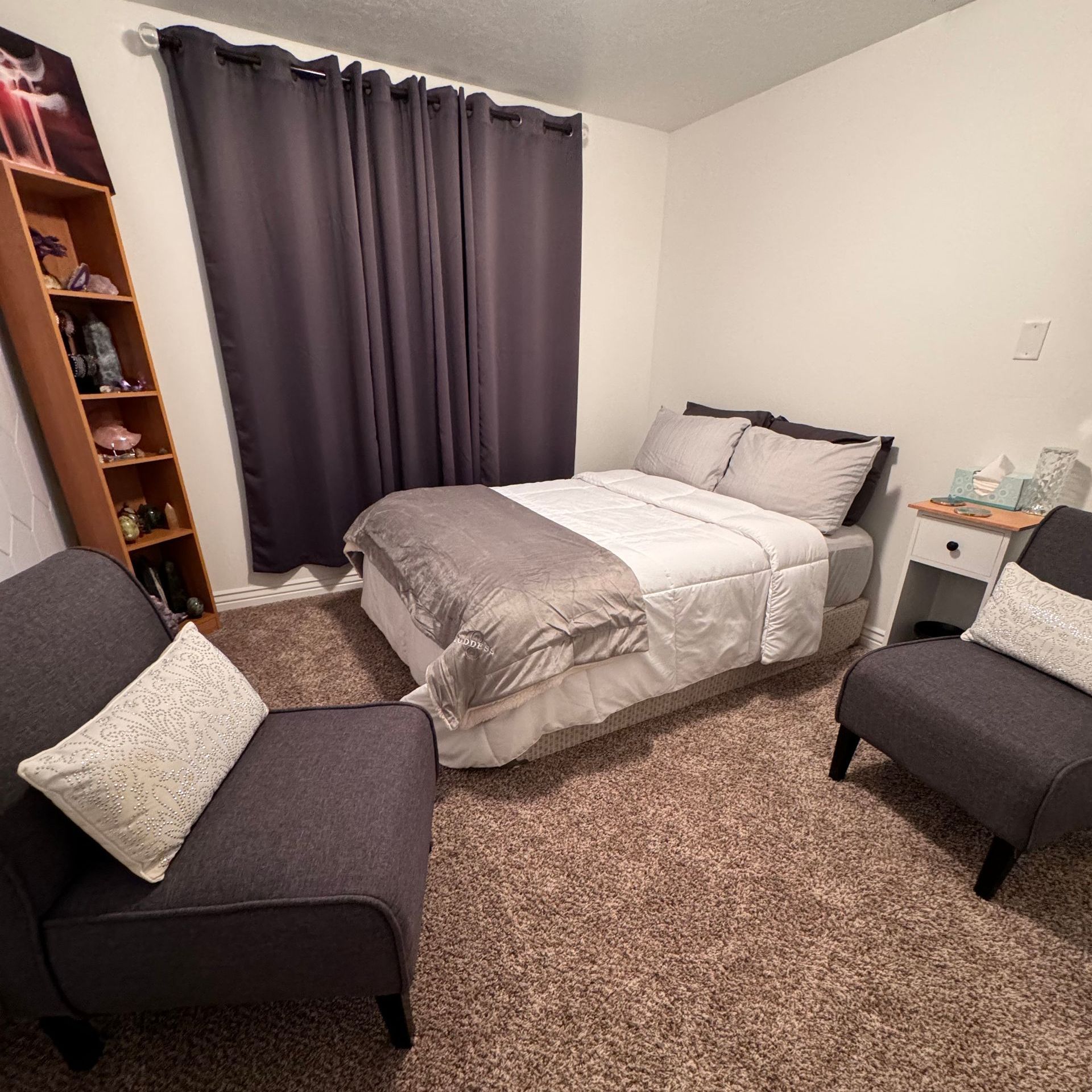 Bedroom with bed, two armchairs, nightstand, bookcase, and gray curtains. Carpeted floor.