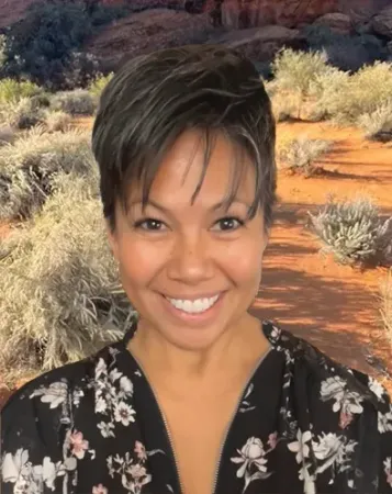 Woman with short dark hair smiles, wearing a floral top, against a desert landscape background.