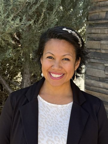 Woman with short dark hair smiles, wearing a floral top, against a desert landscape background.