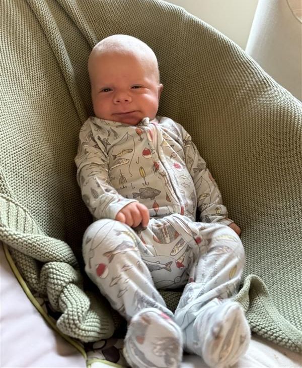 Baby in patterned footie pajamas sits in a green chair, smiling slightly.