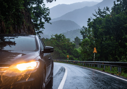 Car driving on a wet mountain road in the rain with headlights on and forested hills in the background.
