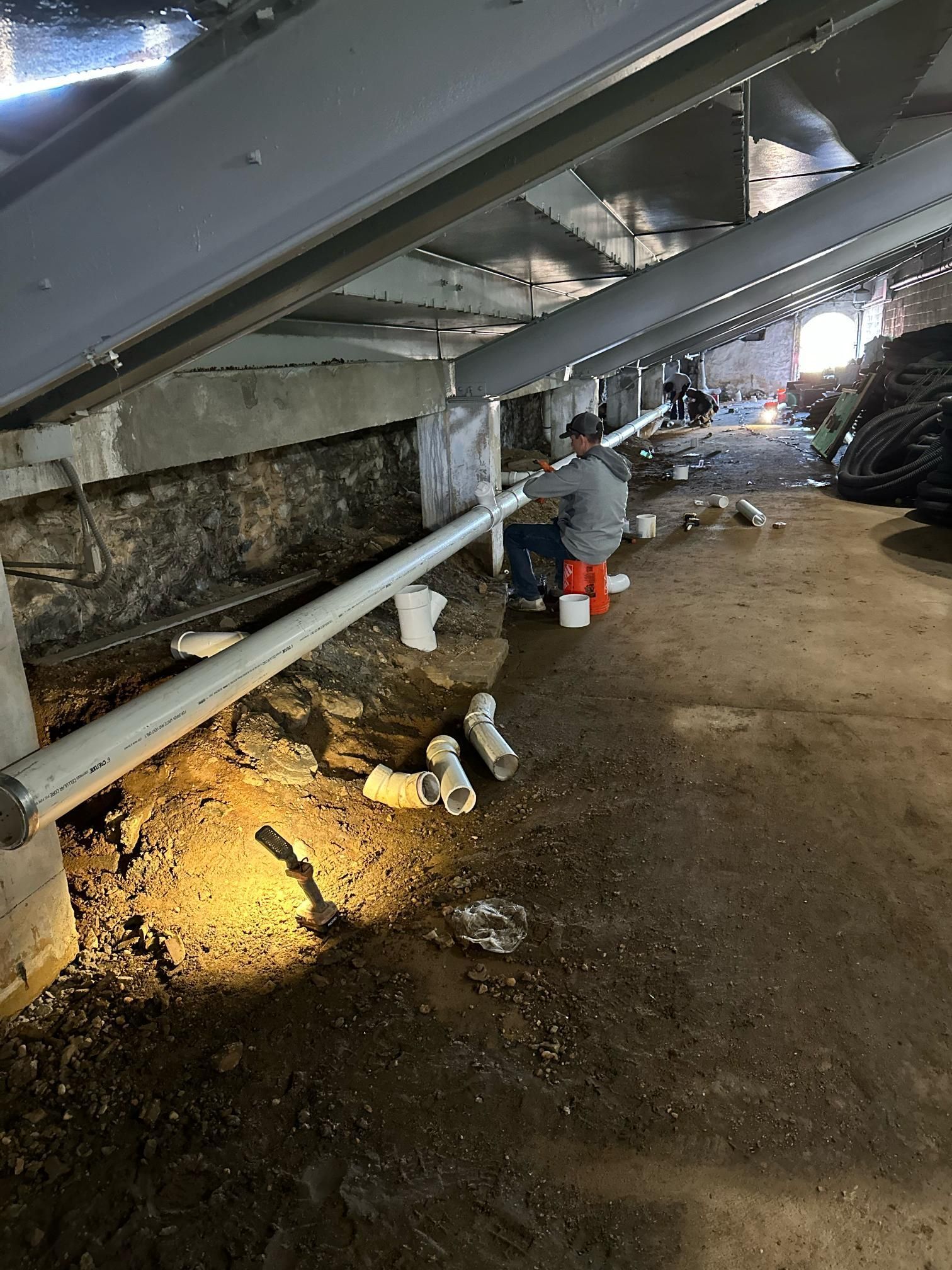 Person installing white pipes in a dim, dirt-floored basement with exposed beams and brick.