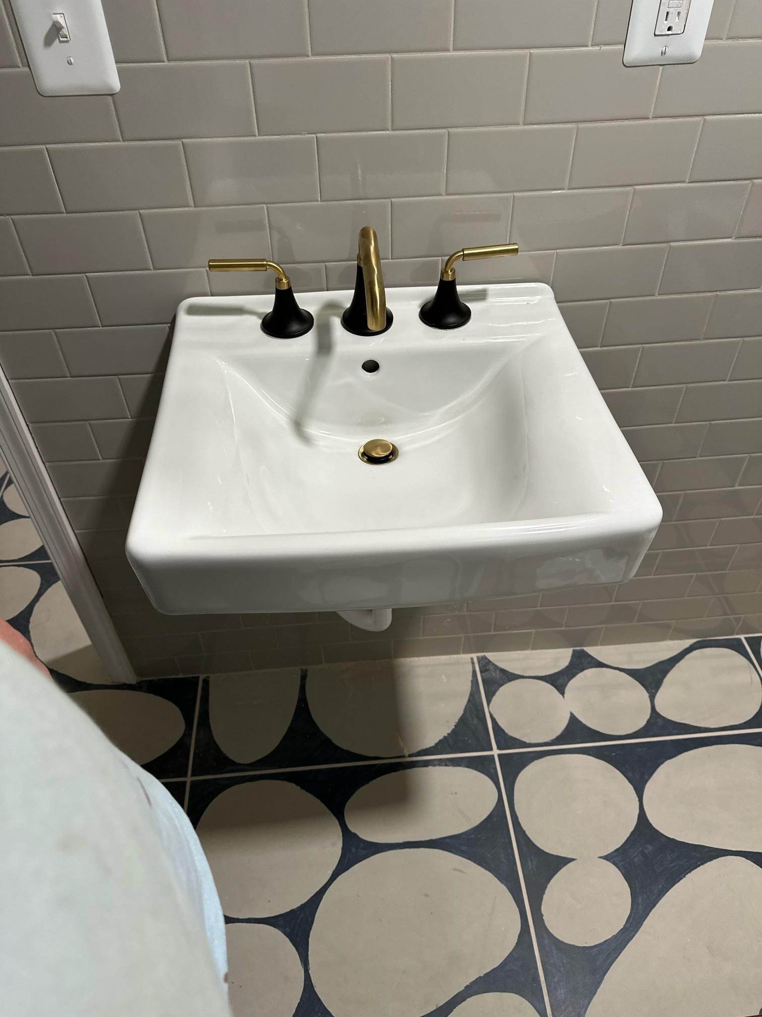 White wall-mounted sink with gold and black fixtures, grey tiled wall, patterned floor.