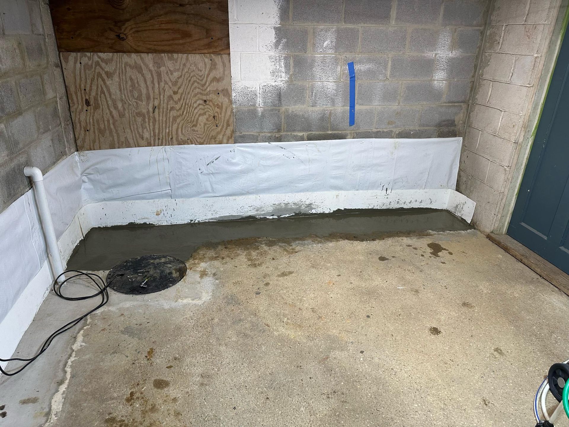 Basement with concrete floor, new cement along the wall with white plastic, sump pump, and a door.