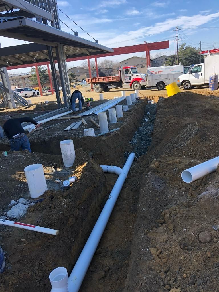 Construction site: workers installing white pipes in a trench. Building framework overhead, red sign, trucks in background.