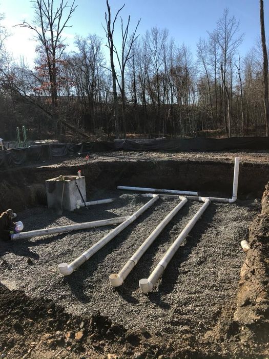 A septic system under construction in a gravel-lined trench, with white pipes and a concrete tank.
