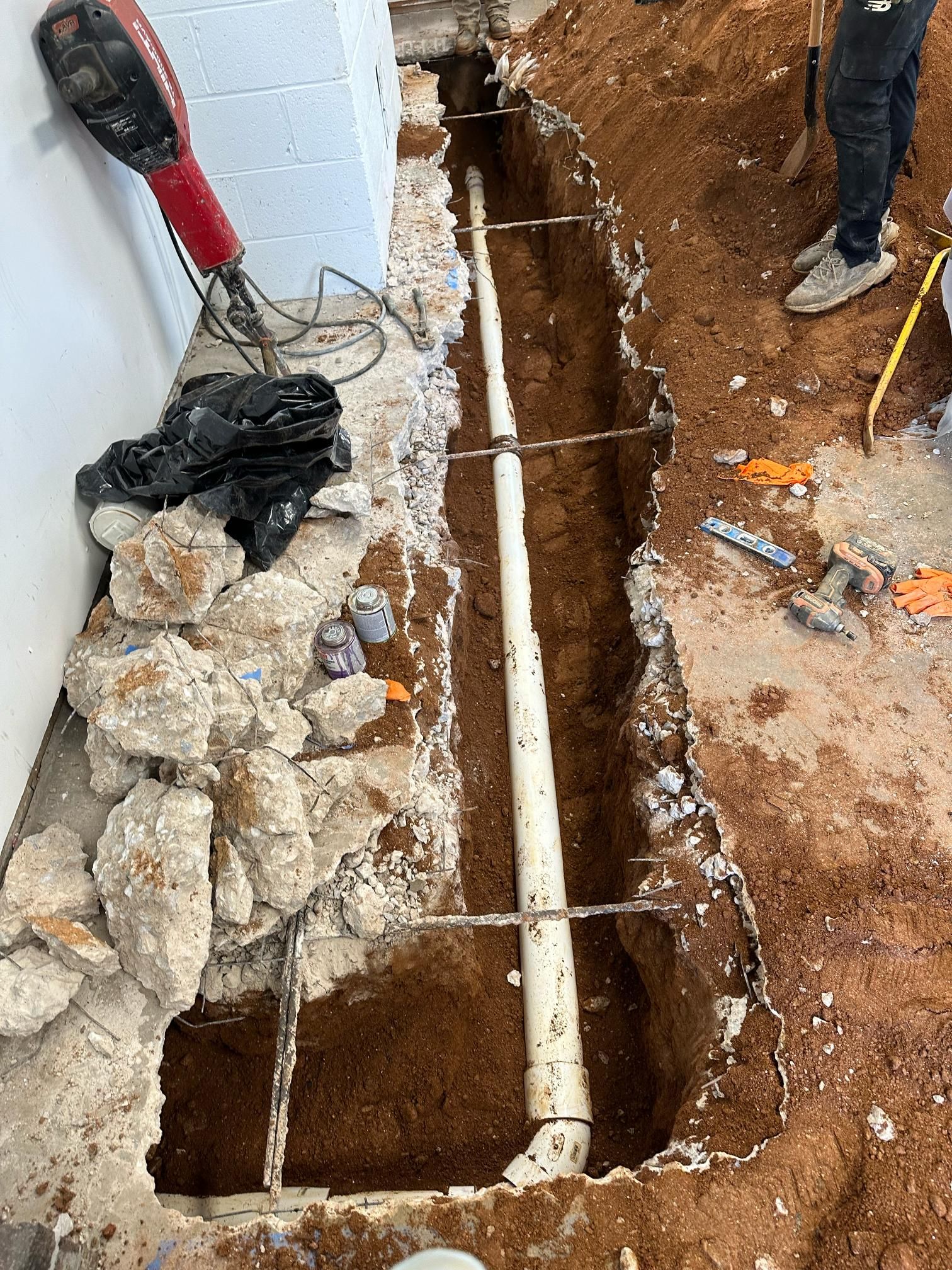 Trench dug alongside a wall, revealing a white PVC pipe. Construction equipment nearby.