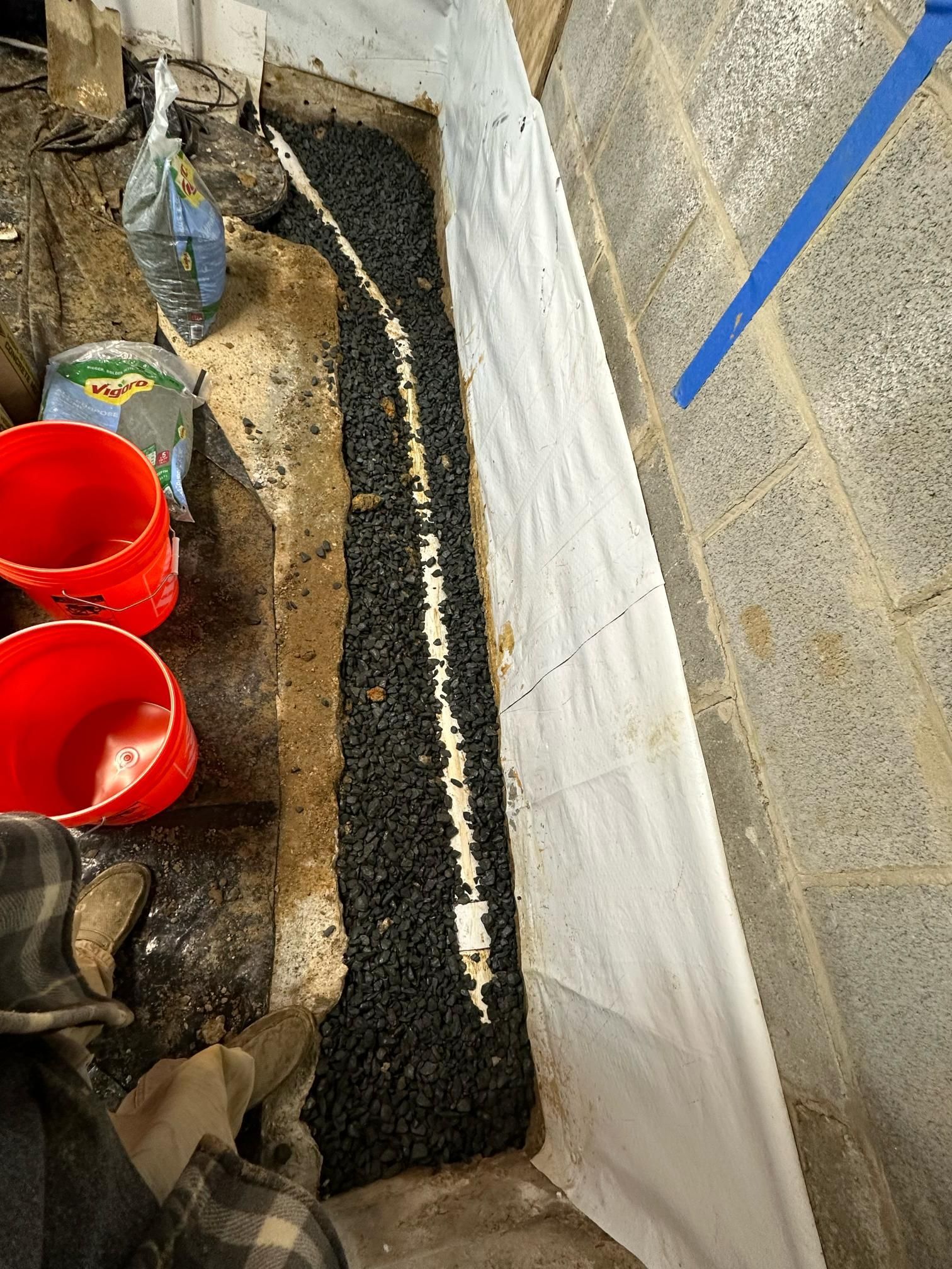 Basement trench filled with black gravel and white pipe, against a concrete wall, with red buckets.