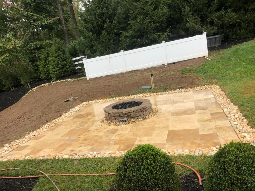 Stone patio with fire pit, bordered by rocks and lawn, sloped backyard with white fence.