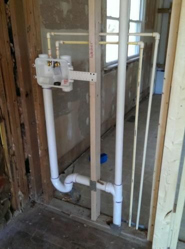 Plumbing rough-in: white PVC pipes and brass fixtures within wooden framing, near a window in a room under renovation.