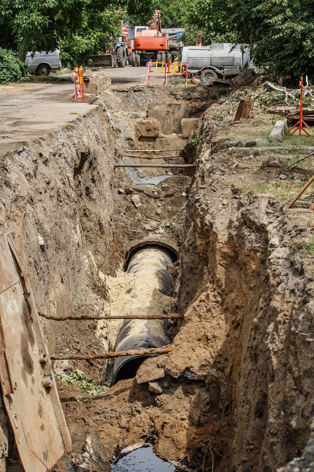 Construction site: trench with a large pipe, equipment and workers in the background.