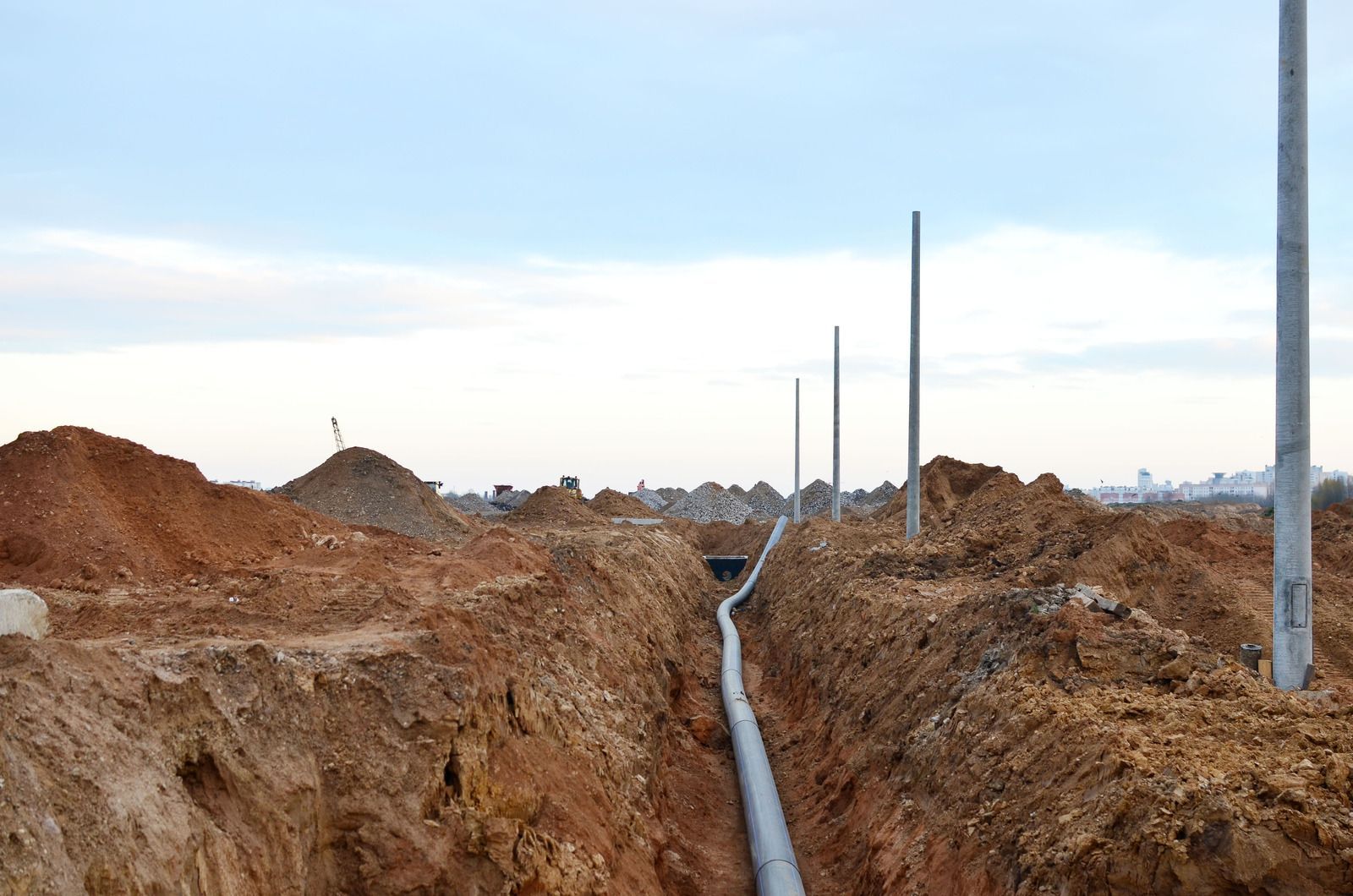 Trench with a pipe running through it, piles of dirt on either side, and utility poles in a construction zone.