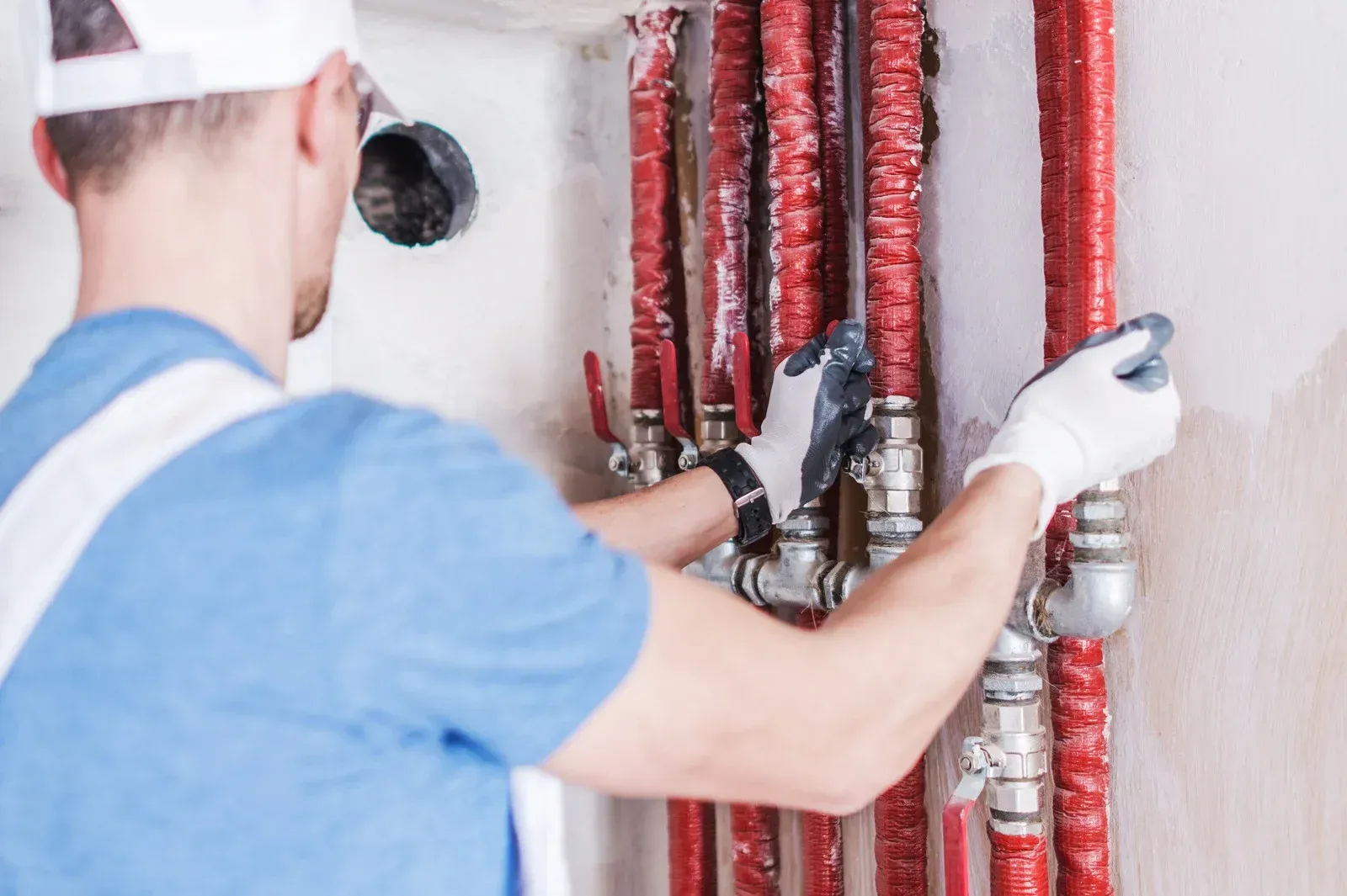 A plumber in a blue shirt and gloves working on red pipes in a white-walled room.