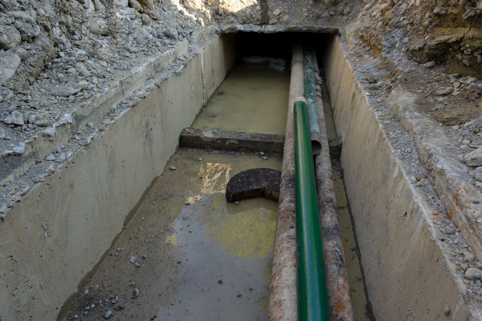 A deep trench in a road reveals a large pipe. Construction equipment and orange cones are visible in the background.
