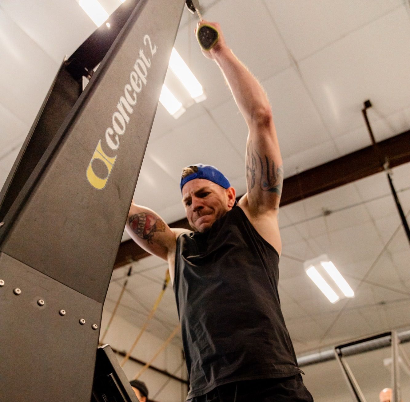 Man exercising on a Concept2 machine, arms raised, indoors, wearing a blue hat, black tank top, and tattoos.