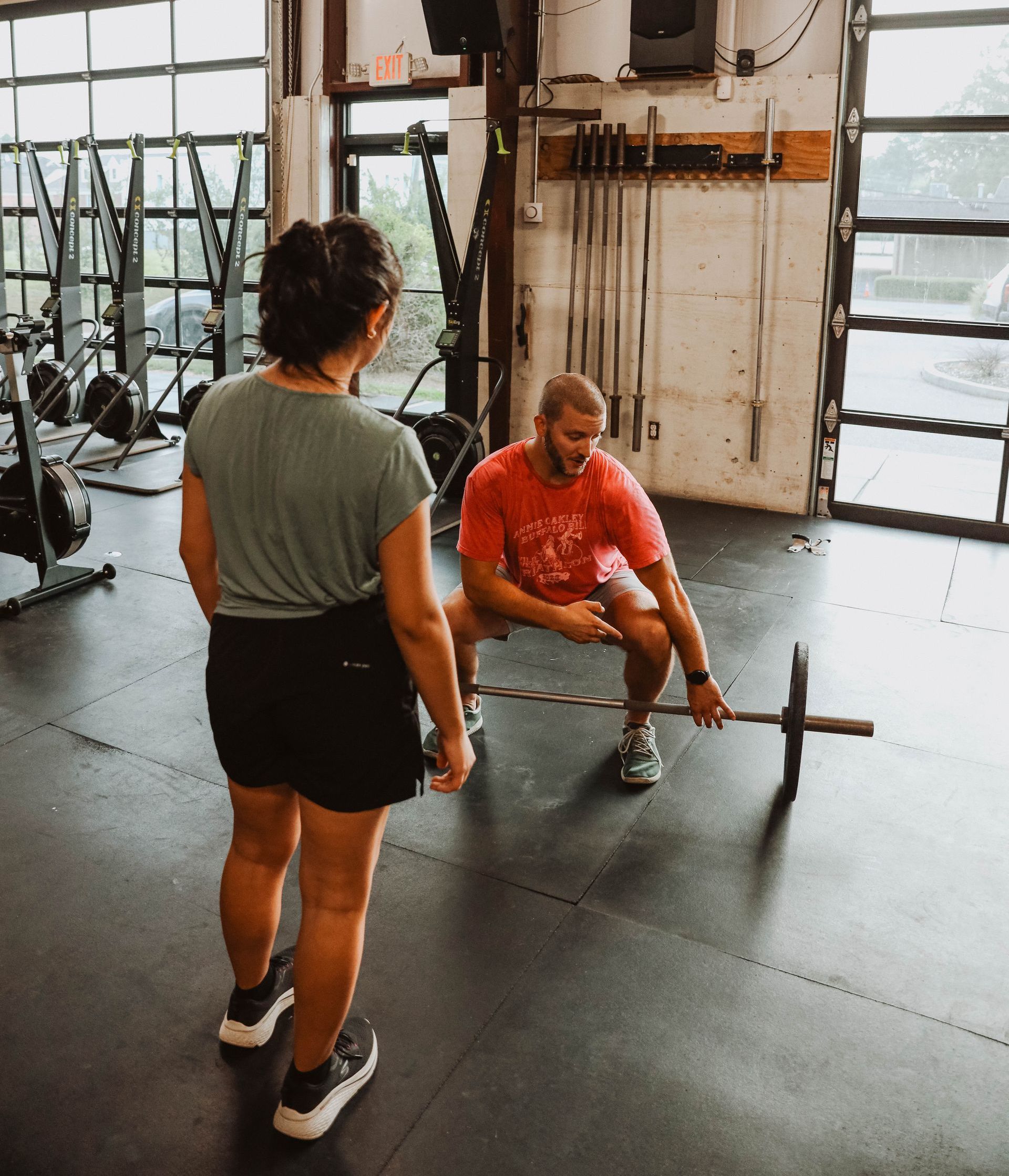 Woman observes man doing a squat with a barbell in a gym. Both are wearing athletic clothing.