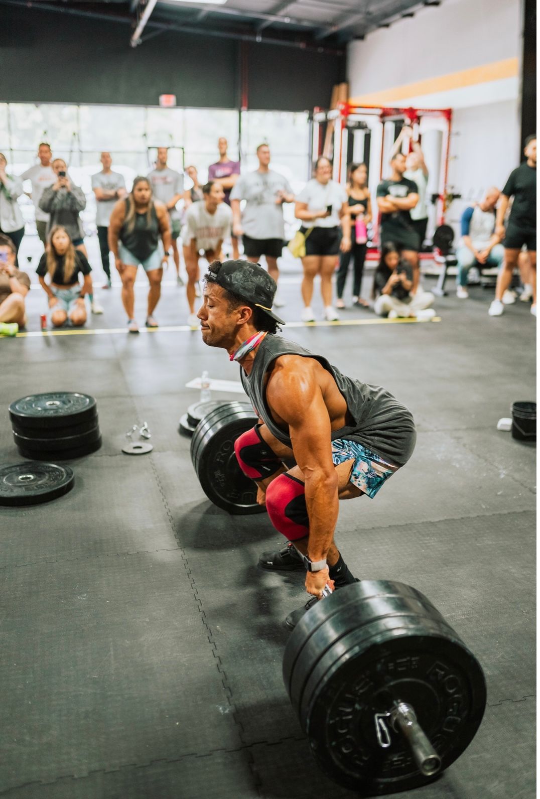 Man in gym lifts a heavy barbell with onlookers. He wears knee sleeves, a tank top, and a hat.