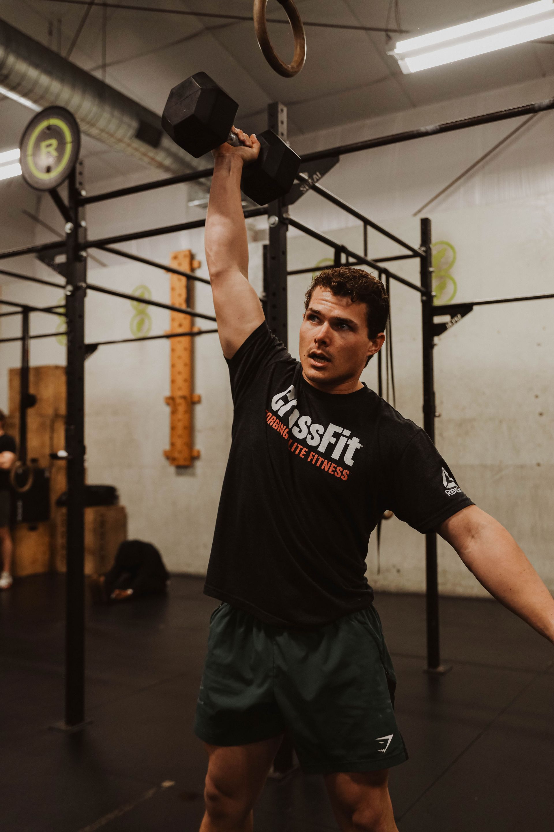 Man in CrossFit gym lifting a dumbbell overhead. He wears a black shirt and green shorts.