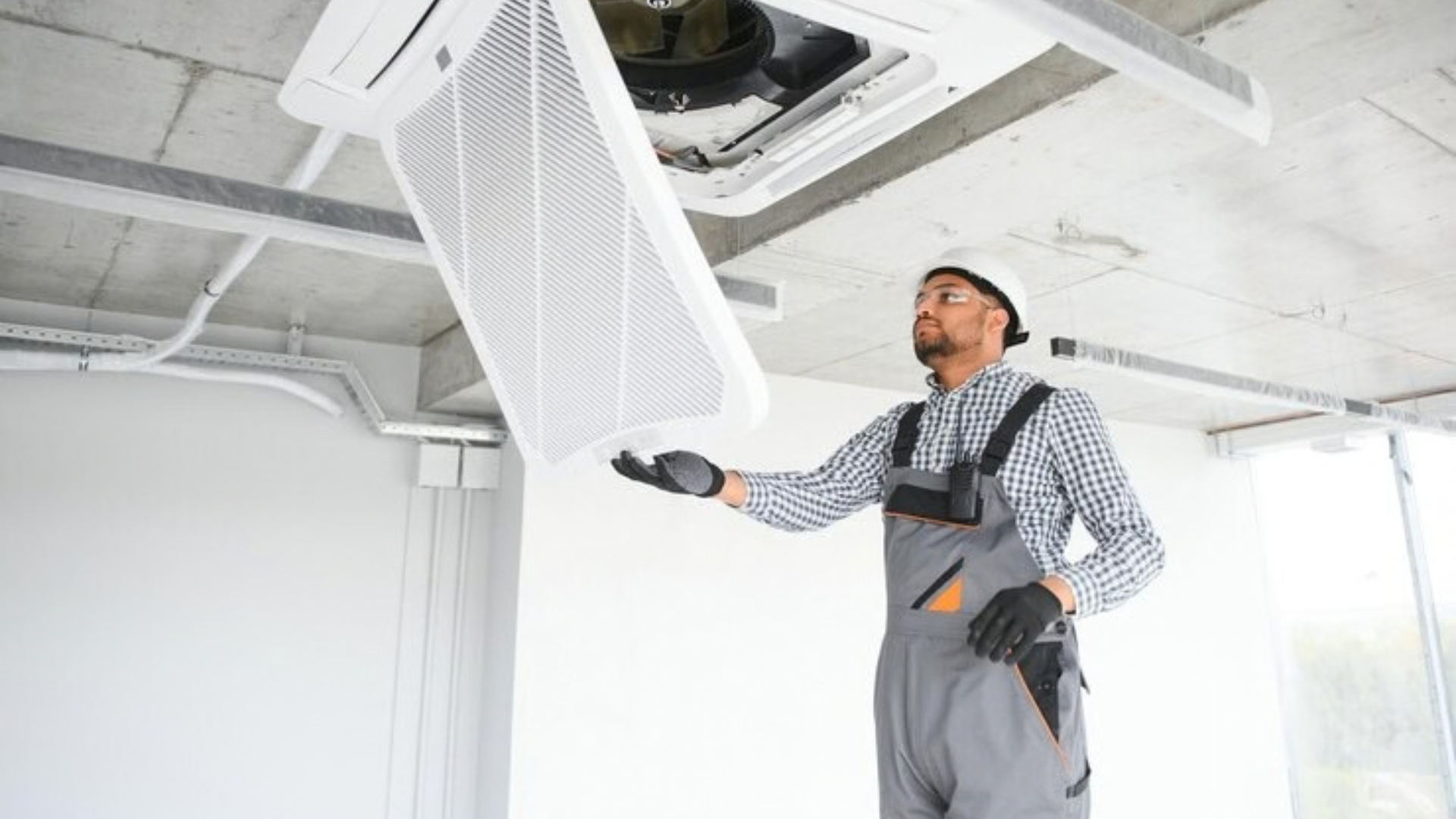A technician in a hard hat and grey overalls repairs a ceiling-mounted air conditioning unit in a room.