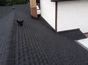 A high-angle view of a dark asphalt shingle roof, featuring a brick chimney, a white wall, and a metal roof vent.