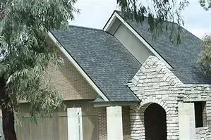 A house featuring a stone entrance, grey shingled roofs, and a textured tan garage exterior under a bright sky.